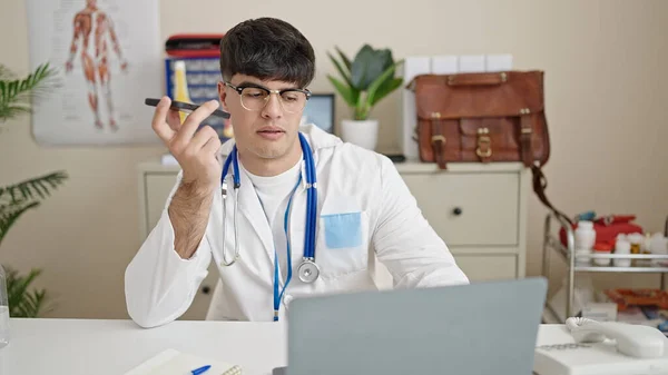 他的年轻医生在诊所用笔记本电脑用智能手机发送语音讯息 — 图库照片