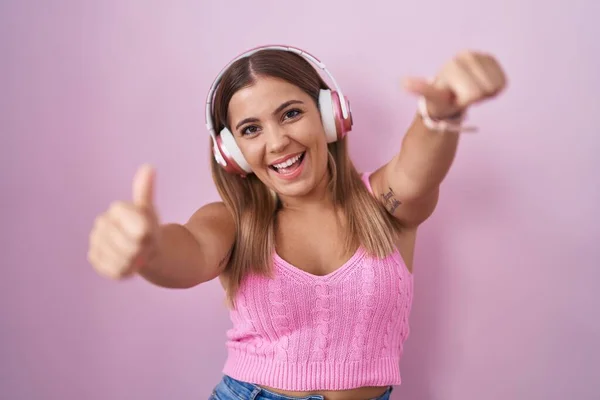Young blonde woman listening to music using headphones approving doing positive gesture with hand, thumbs up smiling and happy for success. winner gesture.