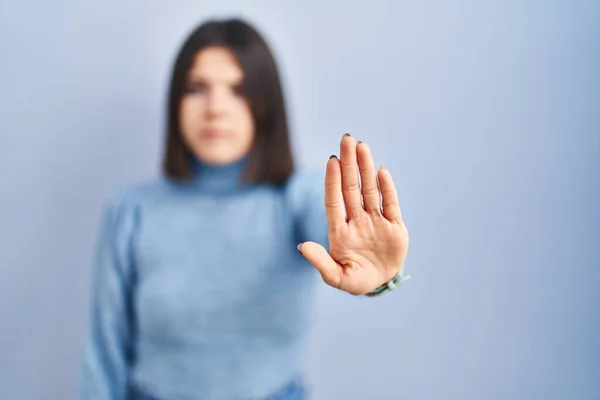 Young hispanic woman standing over blue background doing stop sing with palm of the hand. warning expression with negative and serious gesture on the face.