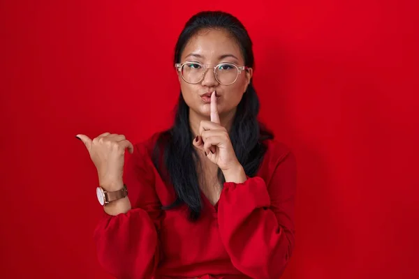 亚洲的年轻女子站在红色的背景上 要求保持安静 手指放在嘴唇上 手放在侧面 沉默和秘密概念 — 图库照片