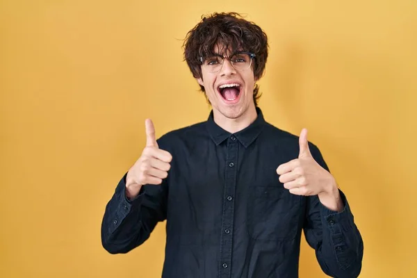 Młody Człowiek Noszący Okulary Żółtym Tle Znak Sukcesu Robi Pozytywny — Zdjęcie stockowe