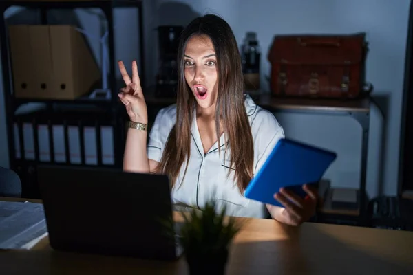 夜のオフィスで働く若いブルネットの女性は 指で勝利のサインをするカメラでウィンクする幸せな顔で笑っています — ストック写真