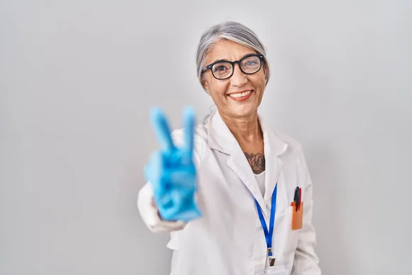 科学者のローブを身に着けている灰色の髪をした中年の女性は 勝利のサインを行う指を示すカメラを見て微笑んだ — ストック写真