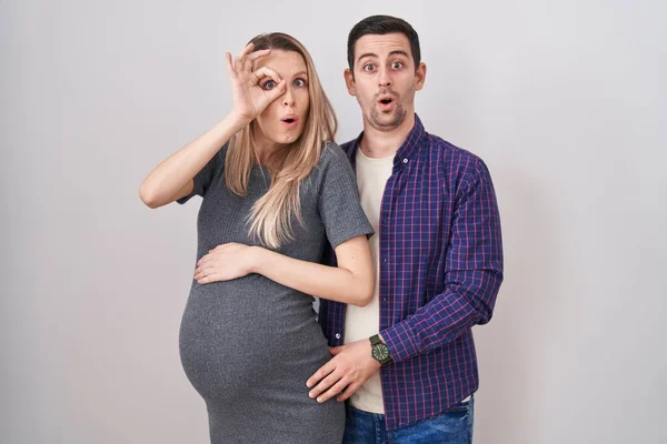若いカップルは 白い背景の上に立っている赤ちゃんを期待し 驚きの顔でショックを受けたOkジェスチャーを行うと 目の指を見て 信じられない表現 — ストック写真