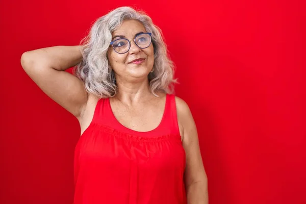 Middle age woman with grey hair standing over red background smiling confident touching hair with hand up gesture, posing attractive and fashionable