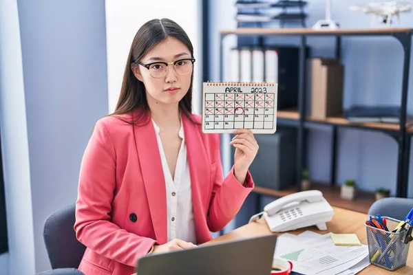 Chinese young woman working at the office holding holidays calendar thinking attitude and sober expression looking self confident