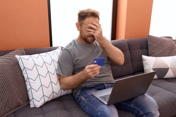 Middle age man using laptop and credit card sitting on sofa at home
