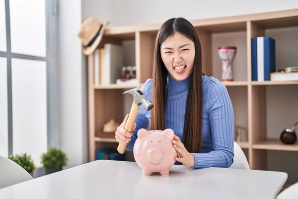 Chinese young woman holding hammer and piggy bank sticking tongue out happy with funny expression.