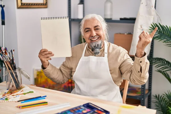 一个白发苍苍的中年男人坐在艺术工作室里 手里拿着笔记本 开心地微笑着 手指手画脚地指向旁边 — 图库照片