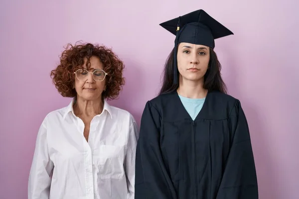 卒業キャップと式のローブを着たヒスパニック系の母親と娘は 顔に深刻な表情でリラックスしました カメラを見ているだけで — ストック写真
