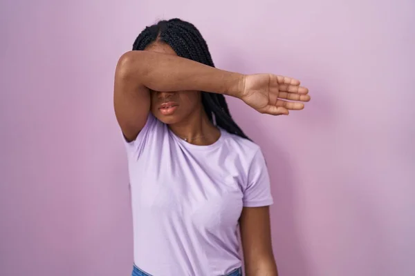 身穿辫子的非洲裔美国女人站在粉色的背景上 用胳膊蒙住眼睛 看起来严肃而忧郁 躲藏和拒绝的概念 — 图库照片
