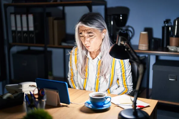 Middle age woman with grey hair working at the office at night with hand on stomach because nausea, painful disease feeling unwell. ache concept.