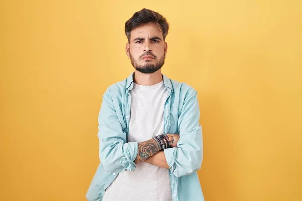 黄色の背景懐疑的で緊張した上に立って入れ墨を持つ若いヒスパニック系の男 交差腕で顔に不承認式 否定的な人 — ストック写真