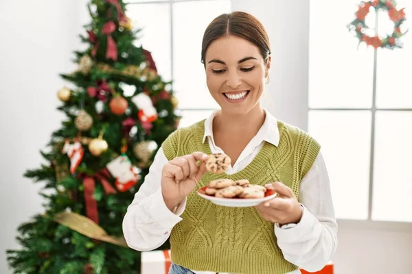 年轻美丽的惊慌失措的女人在家里吃着圣诞树旁的饼干 — 图库照片
