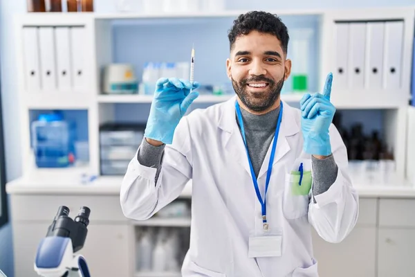 西班牙裔男子 留着胡子 在科学家实验室工作 拿着注射器 高兴地微笑着 手指手画脚地指向旁边 — 图库照片