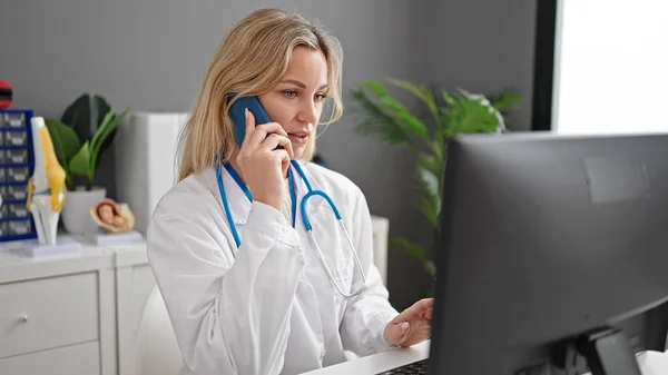 若いですブロンド女性医師使用コンピュータ話して上のスマートフォンでクリニック — ストック写真