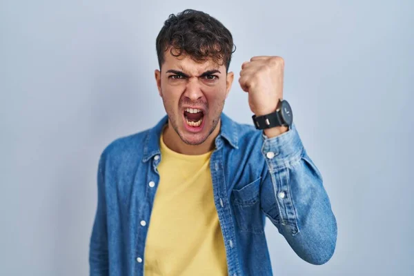 青い背景に立っている若いヒスパニック系の男は怒りで叫んでいる間に怒りと怒りに苛まれ 激しく拳を上げる 怒りと攻撃的な考え方 — ストック写真