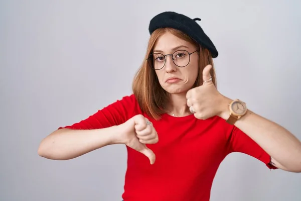 Young redhead woman standing wearing glasses and beret doing thumbs up and down, disagreement and agreement expression. crazy conflict