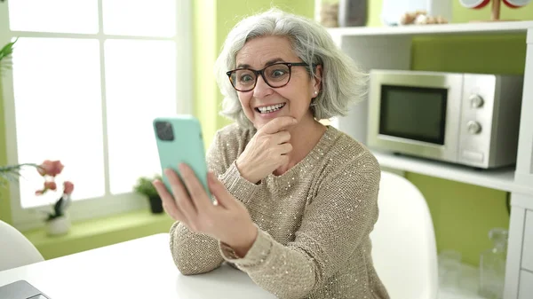 自宅のテーブルに座ってスマートフォンを使用して白髪の中年女性 — ストック写真