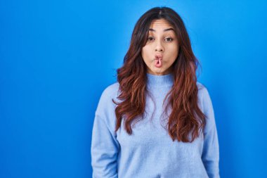 Latin kökenli genç bir kadın mavi arka planda duruyor. Dudaklarıyla balık suratı yapıyor. Çılgınca ve komik bir hareket. Komik ifade. 