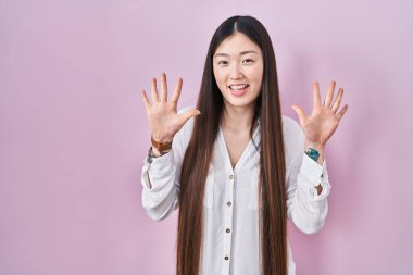 Chinese young woman standing over pink background showing and pointing up with fingers number ten while smiling confident and happy. 