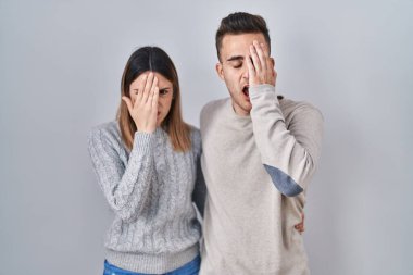 Young hispanic couple standing over white background yawning tired covering half face, eye and mouth with hand. face hurts in pain. 