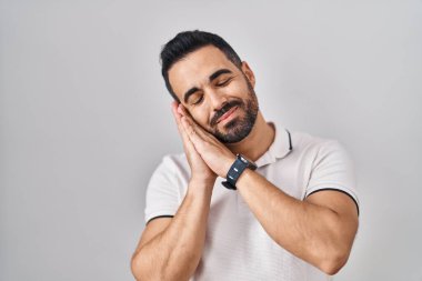 Sakallı, beyaz arka planda günlük giysiler giyen, yorgun, hayal kuran ve elleri ile birlikte poz veren, gözleri kapalı genç İspanyol bir adam.. 