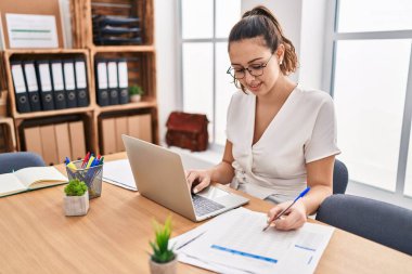 Genç, güzel İspanyol iş kadını. Ofisteki evraklarda dizüstü bilgisayar kullanıyor.