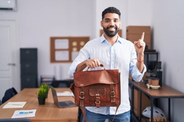 Ofiste çalışan sakallı İspanyol bir adam evrak çantasını tutarken gülümsüyor. 