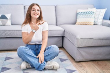 Genç, beyaz bir kadın, oturma odasında yerde mutlu bir şekilde oturuyor. Yüzünde, mutlu ve doğal bir ifadeyle elleri ve parmaklarıyla işaret eden bir gülümsemeyle. 