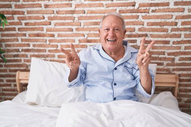 Orta yaşlı, kır saçlı, zafer jesti yapan bir adam yatak odasında oturuyor.