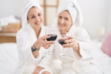 Mother and daughter wearing bathrobe drinking glass of wine at bedroom