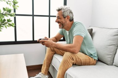 Middle age grey-haired man playing video game sitting on sofa at home