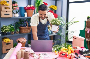 Young hispanic man florist using laptop holding plant pot at florist