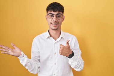 Young hispanic man standing over yellow background showing palm hand and doing ok gesture with thumbs up, smiling happy and cheerful 