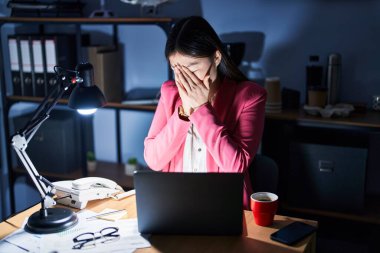 Geceleri ofiste çalışan Çinli genç bir kadın. Ağlarken yüzünü ellerle kapatan üzgün bir ifadeyle. depresyon kavramı. 