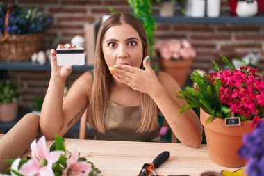 Çiçekçide çalışan genç sarışın kadın elinde kredi kartıyla ağzını kapatıyor, şok geçiriyor ve hata yapmaktan korkuyor. şaşırmış ifade 