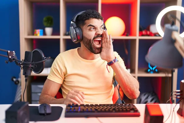 髭を生やしたヒスパニック系の男がヘッドフォンでビデオゲームをプレイし 大声で叫び 口の中で手で横に叫んでいます コミュニケーションの概念 — ストック写真