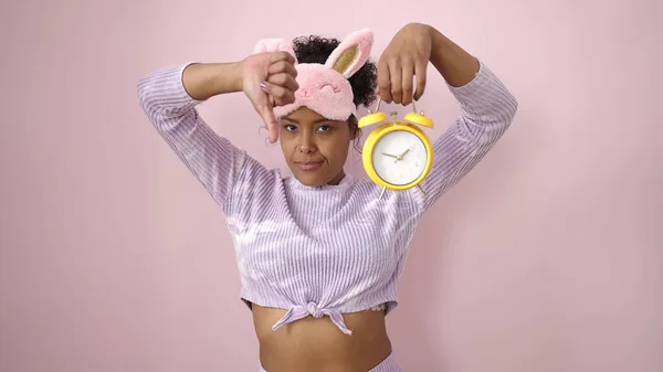 アフリカ系アメリカ人女性身に着けています睡眠マスク保持カップ目覚まし時計とともに親指ダウン隔離されたピンクの背景 — ストック写真