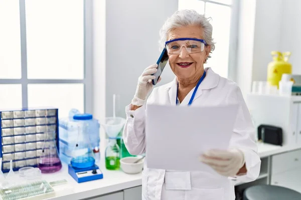 研究室でスマートフォンで話す科学者の制服を着たシニア白髪の女性 — ストック写真