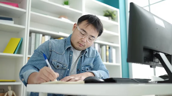 Young chinese man student using computer writing on notebook at library university