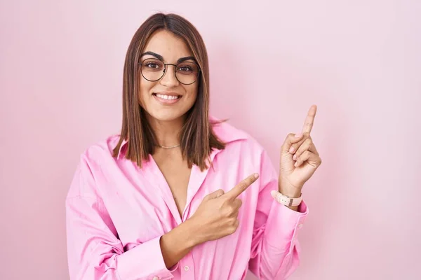 年轻的惊慌失措的女人 戴着眼镜 站在粉色的背景上微笑着 看着摄像机 用双手和手指指向旁边 — 图库照片