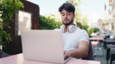 Young hispanic man listening to music sitting on table at coffee shop terrace