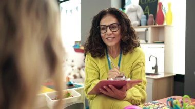 Middle age hispanic woman preschool teacher smiling confident writing on document at kindergarten