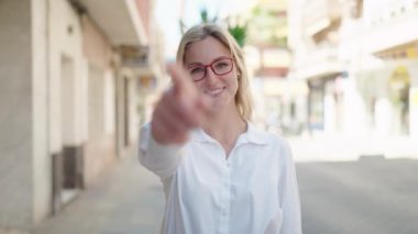 Young blonde woman smiling confident doing coming gesture with finger at street