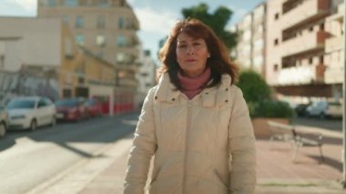 Orta yaşlı kızıl saçlı kadın gülümsüyor. Kollarını kavuşturup caddede duruyor.