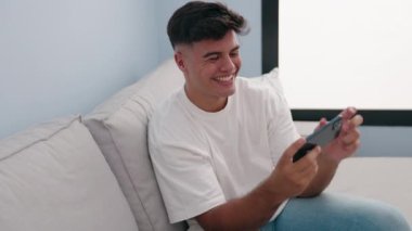 Genç İspanyol adam akıllı telefondan video izliyor. Evdeki kanepede oturuyor.