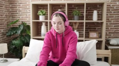 Young blonde woman dancing and listening to music sitting on bed at bedroom