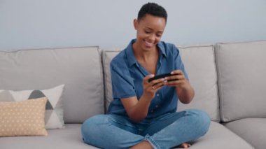 Afro-Amerikalı kadın akıllı telefonuyla evde video oyunu oynuyor.