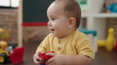 Adorable hispanic baby playing with construction blocks sitting on floor at kindergarten